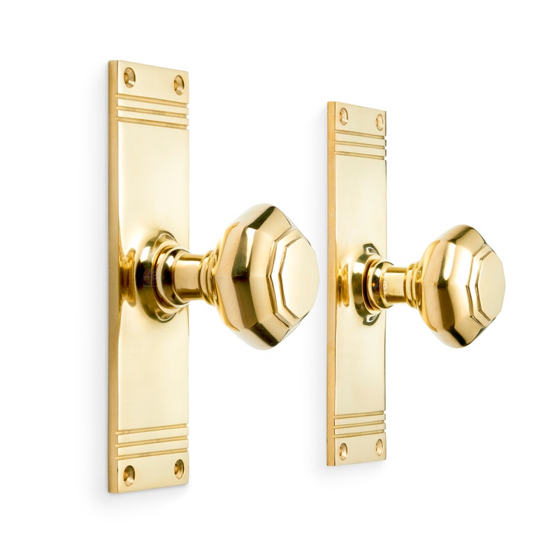 May include: Two polished brass door handles. Each handle has a rectangular backplate with horizontal grooves and a faceted, geometric knob. The handles are mounted on a white surface.