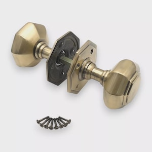 May include: A pair of antique brass door knobs with a faceted design. The set includes two knobs, two square backplates, and a set of screws. The hardware is a warm gold colour and is set against a neutral background.