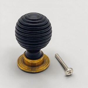 May include: A black, ribbed cabinet knob with a brass base and a silver screw. The knob is spherical with horizontal grooves. The brass base has a circular design. The screw is silver and has a flat head.