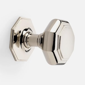 May include: A polished nickel door knob. It features an octagonal backplate and a faceted handle. The hardware has a reflective surface and a classic design, suitable for interior doors. The knob is mounted on a white surface.