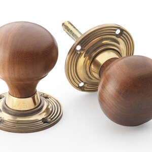May include: Two wooden door knobs with brass detailing. The knobs are a rich brown colour and spherical. The brass base features a concentric circle design. One knob is disassembled, revealing the threaded rod.