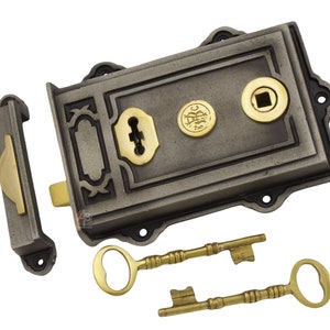 May include: A traditional door lock set in a grey metal finish with brass details. The set includes the lock mechanism, a strike plate, and two decorative keys. The lock features a keyhole and a circular emblem.