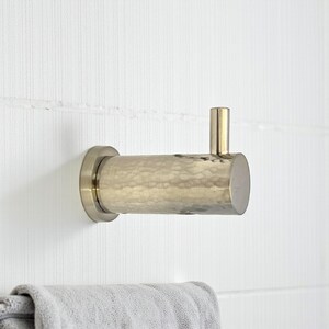 May include: A brushed nickel wall hook with a hammered texture. The hook has a cylindrical body and a small, vertical post. It is mounted on a white, textured wall. A grey towel hangs below.