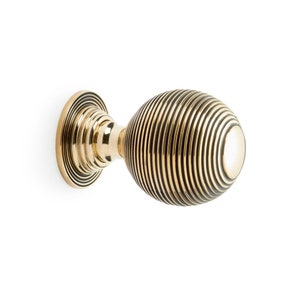 May include: A gold-coloured door knob with a round, ribbed design. The knob is attached to a circular base and has a spherical shape. The ribbed pattern creates a textured appearance. The hardware is set against a white background.