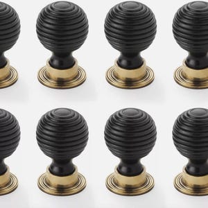May include: Eight black cabinet knobs with a ribbed, spherical design. Each knob has a brass-coloured base and stem. The knobs are arranged in two rows of four against a white background. These knobs are suitable for furniture and home decor.
