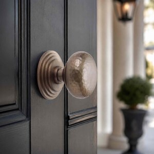 May include: A round, hammered metal door knob with a brushed finish, featuring a circular stepped backplate. The knob is mounted on a dark, paneled wooden door, showcasing a textured surface and a metallic sheen that reflects light across its surface.