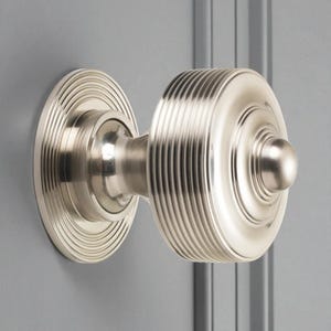May include: A brushed nickel door knob with a round, ribbed handle and a circular backplate. The door knob is mounted on a grey door, showcasing its elegant design and metallic finish. This hardware is suitable for interior doors.