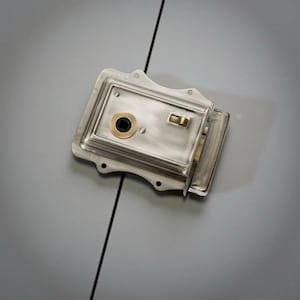 May include: A silver-coloured, rectangular door lock with a brass-coloured circular keyhole and a rectangular latch. The lock has a vintage aesthetic, with visible screw holes and a slightly distressed finish. The lock is designed for security and is likely for a door.