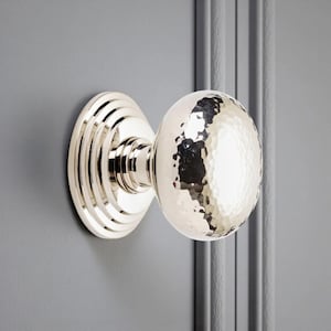 May include: A polished nickel door knob with a hammered texture on the round handle. The knob is mounted on a grey door, showcasing a classic design. The backplate has a layered, circular design.
