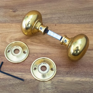 May include: Brass door hardware set comprising two oval doorknobs, two circular backplates, and a small Allen key. The doorknobs are linked by a metal spindle. The items are arranged on a wooden surface.