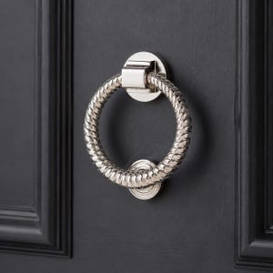 May include: A polished silver door knocker featuring a circular handle with a rope-like texture. It's mounted on a dark grey door. The handle is connected to a round base with a square fitting.