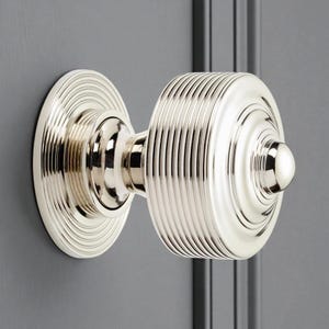 May include: A polished nickel door knob. The circular backplate and cylindrical handle have concentric ridges. The door furniture is mounted on a grey door. The door knob is a classic design.
