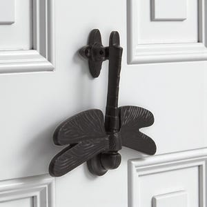May include: A dark bronze dragonfly door knocker fixed to a white door. The knocker has detailed wings and a long, slender body. The dragonfly design adds a decorative touch to the door.