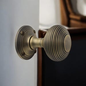 May include: Antique brass door knob with a round, ribbed design. The knob is mounted on a white surface, with a matching circular backplate. The design features a classic, vintage aesthetic, suitable for interior doors.