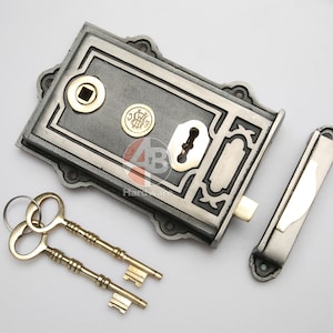 May include: A vintage door lock set in a silver finish, comprising a rectangular lock body, a separate latch, and two gold keys. The lock features ornate detailing and a circular emblem, typical of antique designs.