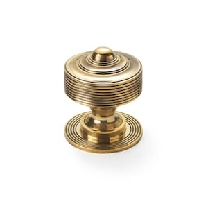 May include: A brass cabinet knob with a tiered, circular design. The knob has a small, rounded top and a base with concentric grooves. The body of the knob features horizontal grooves, giving it a textured look. The hardware is set against a white background.