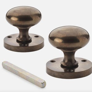 May include: Two antique bronze door knobs with a matching spindle. The knobs have a rounded shape and sit on a circular base with screw holes. The spindle is a long, hexagonal metal rod.