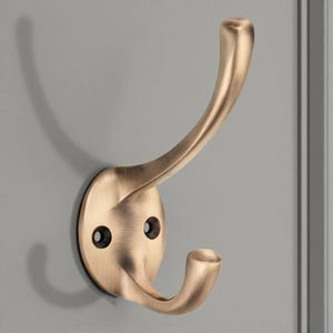 May include: A bronze-coloured wall hook with a double-pronged design. The hook has a curved upper arm and a lower arm, both ending in rounded tips. The hook is mounted on a circular base with two screw holes.
