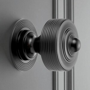 May include: A black door knob with a circular design. The knob features a ridged, cylindrical center and a circular backplate with concentric grooves. The hardware is mounted on a grey surface.
