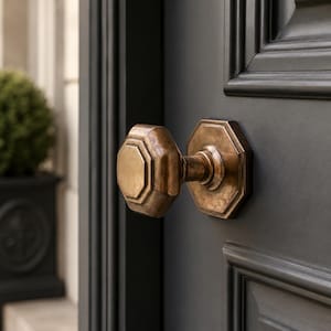 May include: An octagonal brass door knob with a weathered, antique finish mounted on a dark charcoal grey panelled door. The hardware features a matching octagonal backplate and a central stem connecting the knob to the door surface.