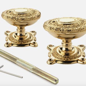 May include: Two decorative gold door knobs with detailed floral patterns. The knobs have ornate bases and are shown with a threaded rod and an Allen key. The hardware is likely for a cabinet or door.