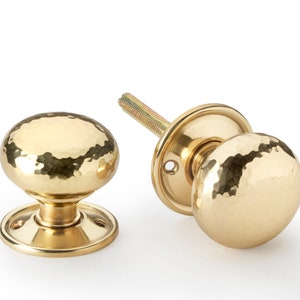May include: Two brass door knobs with a hammered texture. The knobs are round with a circular base and a threaded spindle. The finish is a shiny gold colour. The door knobs are set against a white background.