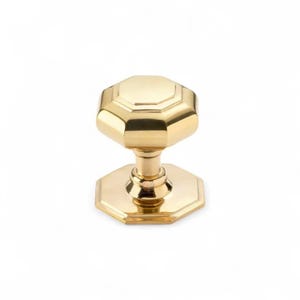 May include: A polished gold door knob with an octagonal design. The knob sits on a matching octagonal base. The metal hardware is suitable for interior doors. The knob's design is symmetrical.