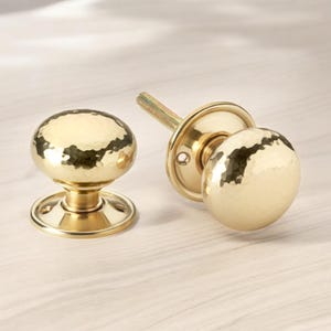 May include: Two gold-coloured door knobs with a hammered texture. One knob is attached to a round backplate with a threaded rod, while the other is separate. The knobs are round with a hammered surface and sit on a round base.