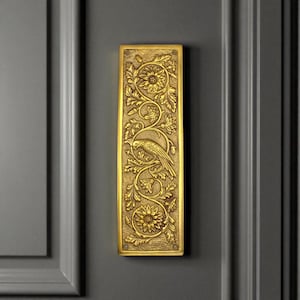 May include: A rectangular, gold-coloured door plate with an embossed floral and bird design. The decorative plate features a bird perched on a branch, surrounded by flowers and leaves. The plate is mounted on a grey door.