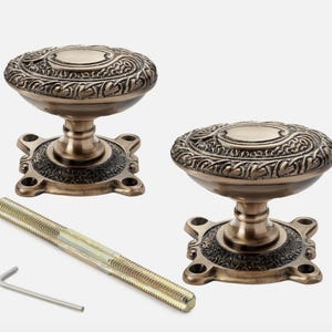 May include: Two antique bronze door knobs with detailed patterns. Each knob features a decorative top and a square base. A long screw and an Allen key are also included.