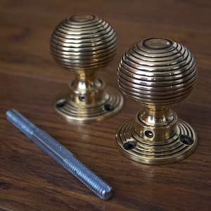 May include: A pair of antique brass door knobs with a spiral design and a silver threaded rod on a wooden surface. The knobs have a round base with three screw holes.