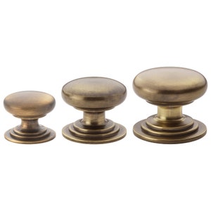 May include: Three antique brass cabinet knobs in varying sizes. Each knob has a rounded top and a decorative base with concentric rings. The knobs are displayed against a white background, highlighting their aged, metallic finish.