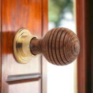 May include: A wooden door knob with a brass base and a grooved, spherical wooden handle. The knob is attached to a wooden door. The background shows a blurred view of a window and foliage.