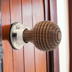 May include: A wooden door knob with a grooved, spherical design. The knob is a dark brown colour and is attached to a silver-coloured metal base. The door is made of vertical wooden planks.