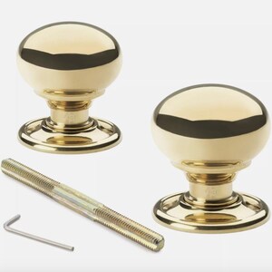 May include: Two polished brass door knobs with a round, dome-shaped design. The knobs sit on a circular base and are accompanied by a threaded rod and an Allen key. The hardware is designed for doors and cabinets.