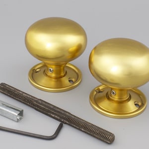 May include: Two gold-coloured door knobs with a rounded design and matching base plates. Included are a threaded rod, a metal bracket, and an Allen key. The hardware is new and ready for installation.