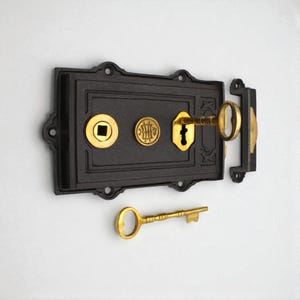 May include: A black antique lock with gold detailing. The lock is rectangular with decorative elements and a keyhole. A gold key is inserted, and another key lies below. The lock is likely for a door or cabinet, a classic design.