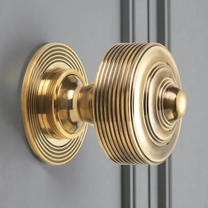 May include: A brass door knob with a circular design. The knob features a round, ribbed centre and a circular base with concentric grooves. The hardware is mounted on a grey door.