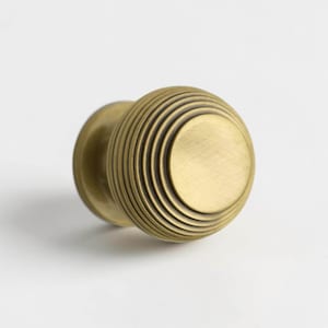 May include: A brass door knob with a circular, ribbed design. The knob has a circular face and a cylindrical base. The ribbed detailing creates a textured, vintage aesthetic. The hardware is likely for a door or cabinet.