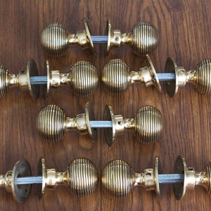May include: Six vintage brass door knobs with a ribbed design, arranged on a wooden surface. Each knob has a round shape and is connected by a metal rod. The knobs are a warm gold colour.