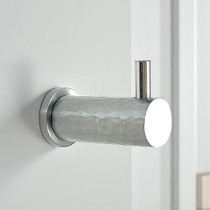 May include: A silver-coloured, hammered metal hook fixed to a white wall. The hook features a cylindrical body with a textured surface and a circular base. A vertical, cylindrical post extends from the end.