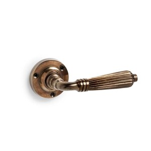 May include: A bronze door handle with a circular backplate and a ribbed lever. The handle is made of metal and has a vintage appearance. The backplate has four screw holes.