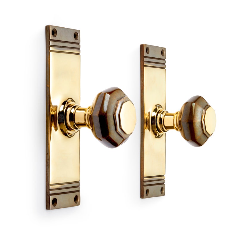 May include: A pair of brass door handles with a rectangular backplate and a decorative, faceted knob. The knobs have a brown and gold color scheme, and the backplates have a two-tone design.