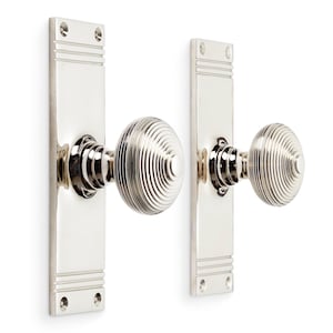 May include: A pair of polished nickel door knobs with rectangular backplates. The knobs feature a circular, ridged pattern. The backplates are rectangular with horizontal grooves at the top and bottom, and screw holes.