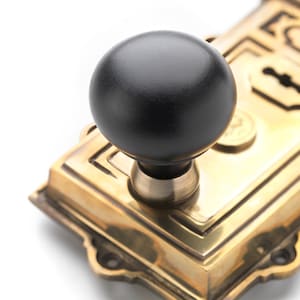 May include: Close-up of a vintage door lock with a black round knob and a brass-coloured metal plate. The lock features a keyhole and decorative detailing, suggesting a classic or antique style.