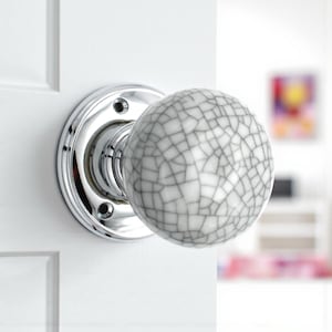 May include: A chrome door knob with a white and grey crackle-patterned ceramic sphere. The knob is mounted on a white door. The door knob is a decorative hardware element.