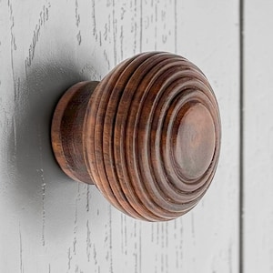 May include: A close-up of a wooden cabinet knob with a circular, grooved design. The knob is a warm brown colour and is mounted on a light grey surface.