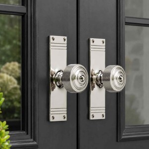 May include: A pair of polished silver-toned metal door knobs with matching rectangular backplates. Each knob features a circular, ribbed design with a smooth centre point. The hardware is mounted onto a dark, matte black double door frame.