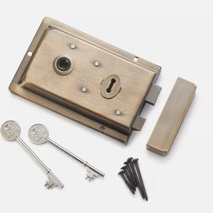May include: A bronze-coloured door lock set, comprising the lock body, a strike plate, two decorative keys, and screws. The lock has an aged appearance with screw holes and a keyhole. The keys have a circular design on the key head.
