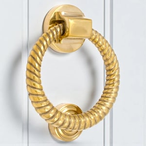 May include: A gold door knocker with a circular, rope-style handle. The knocker is mounted on a white door and has a round base and a square top piece. The design is classic and elegant, suitable for any front door.